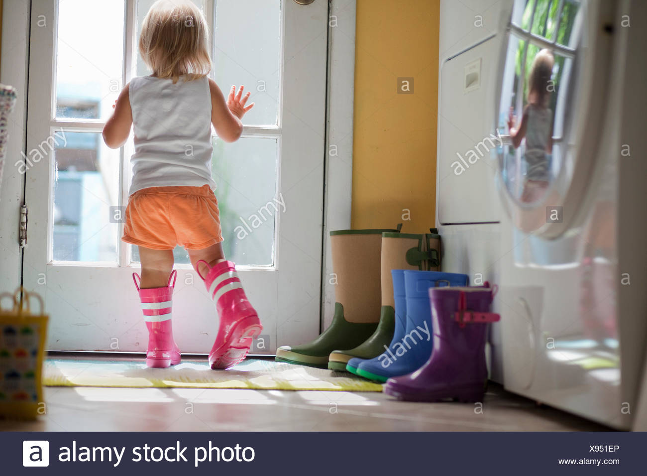 Female Toddler Wearing Rubber Boots Looking Out Of Back Door