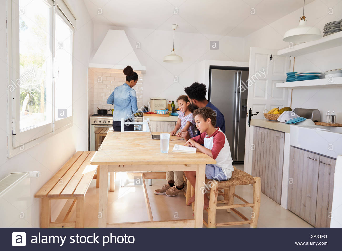 Dad Sitting With Kids At Kitchen Table While Mum Cooks Stock Photo