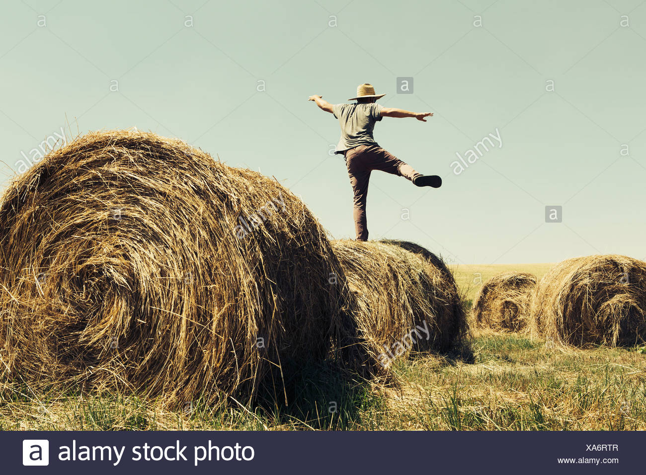 Back view of a man balancing on one leg on top of a hay bale. Stock Photo