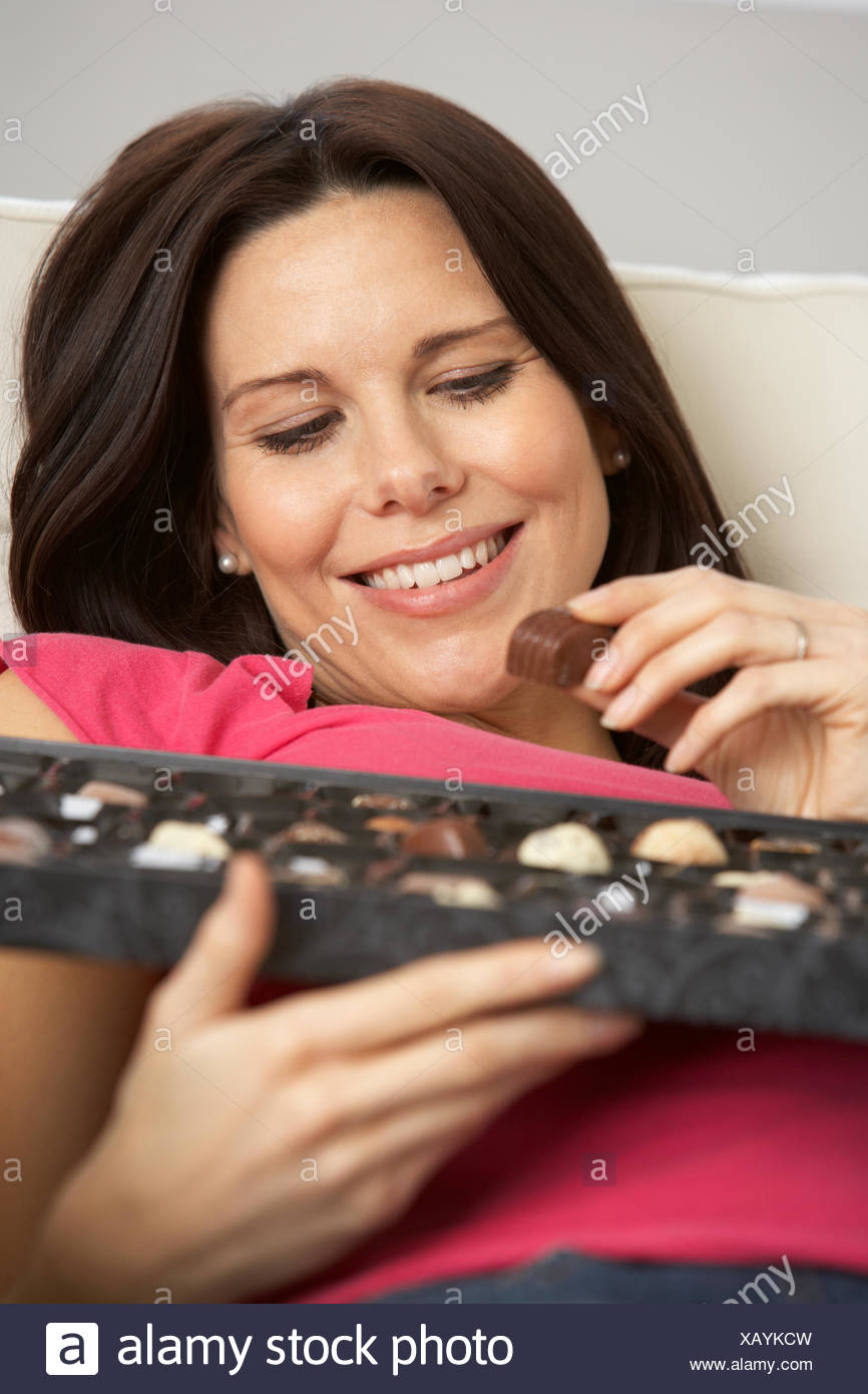 Pregnant Woman Eating Box Of Chocolates Sitting On Sofa At Home