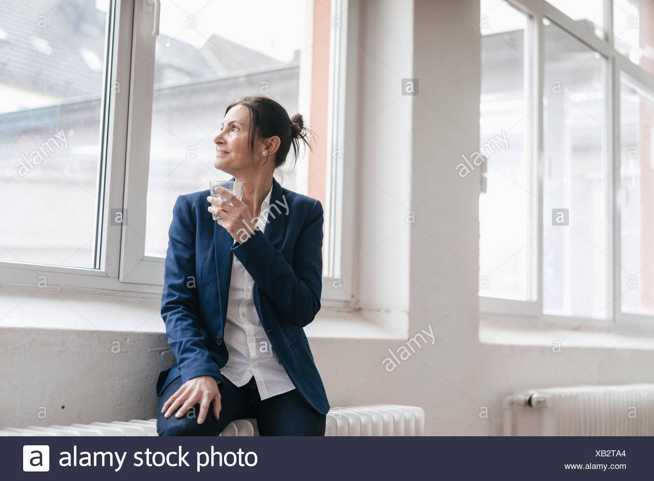Businesswoman with glass of water sitting on radiator in a loft looking  through window Stock Photo - Alamy