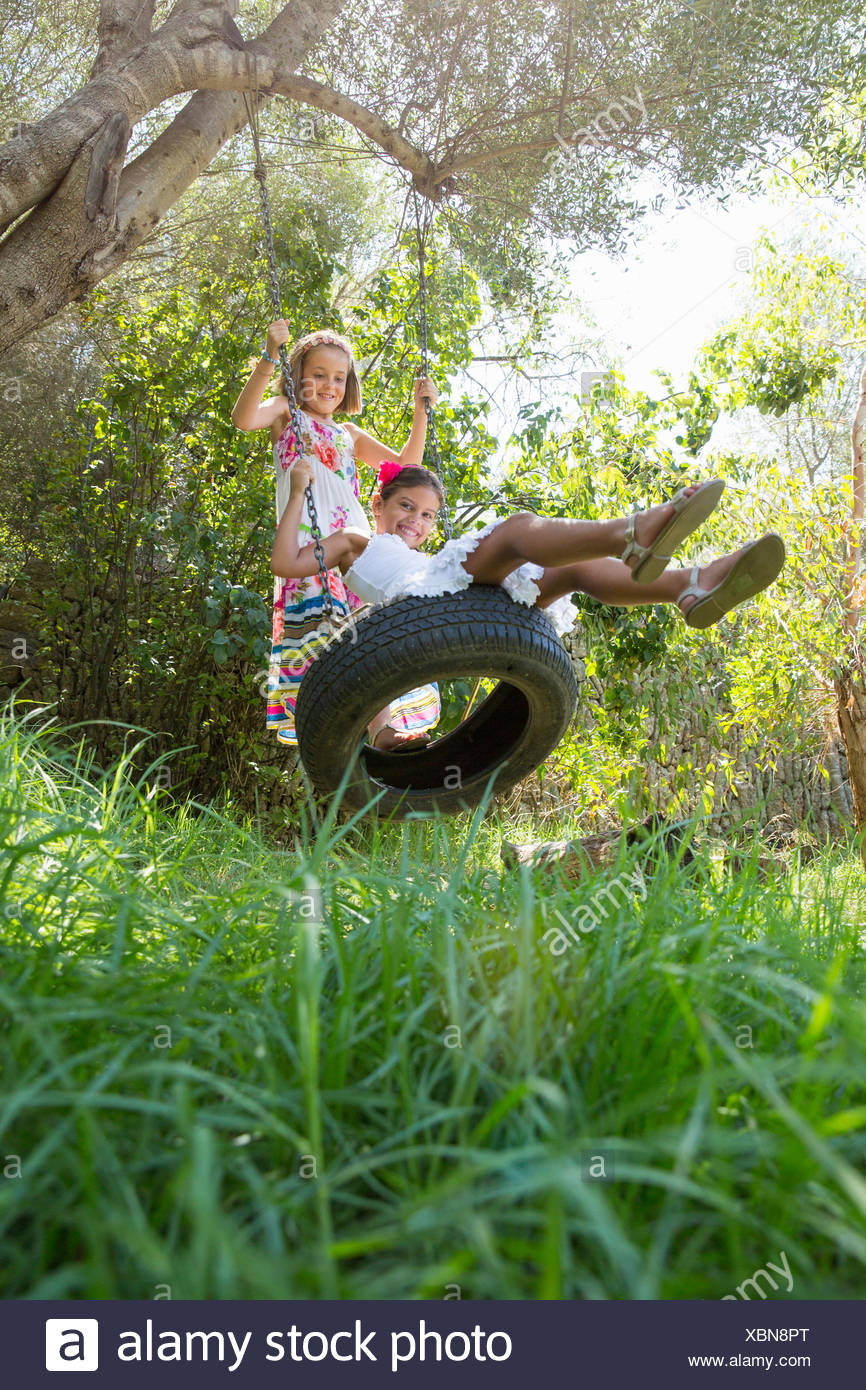 Two Girls Sitting And Standing On Tree Swing In Garden Stock