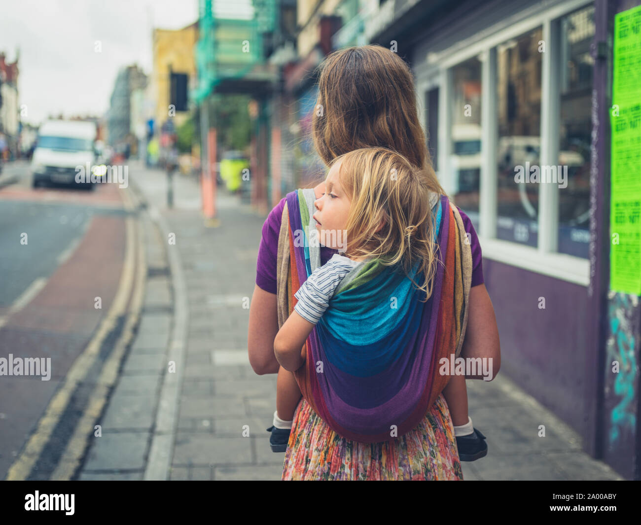 Eine junge tausendjährigen Mutter ist zu Fuß auf der Straße mit ihr Kleinkind zu ihr zurück in einer Schlinge befestigt Stockfoto