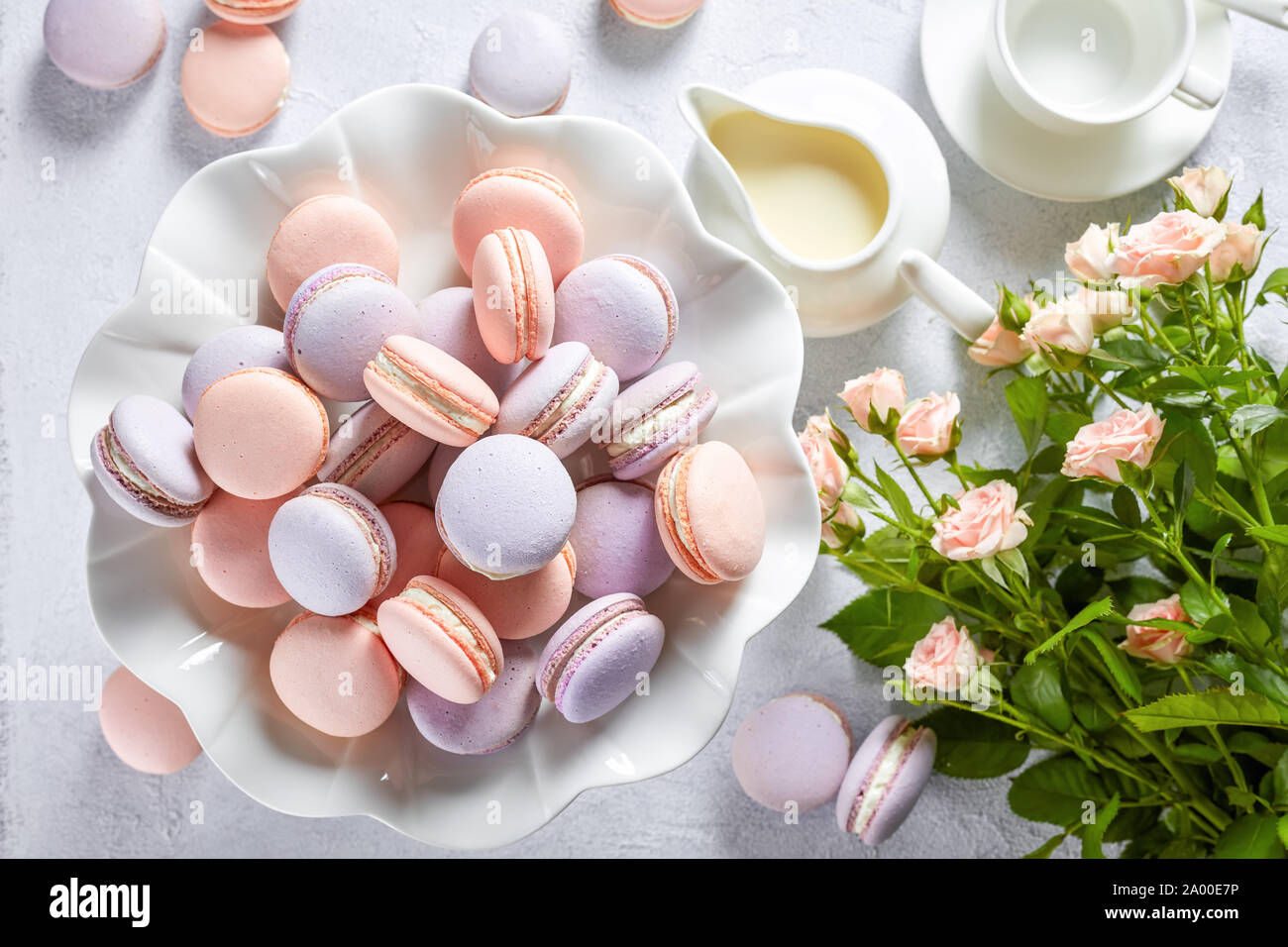 Nahaufnahme der französische Macarons auf einem Porzellan Kuchen stehen und einige auf weißem Beton Tabelle mit Strauß mit frischen Blumen, Kaffee Tasse und Rahmtopf, horiz. Stockfoto