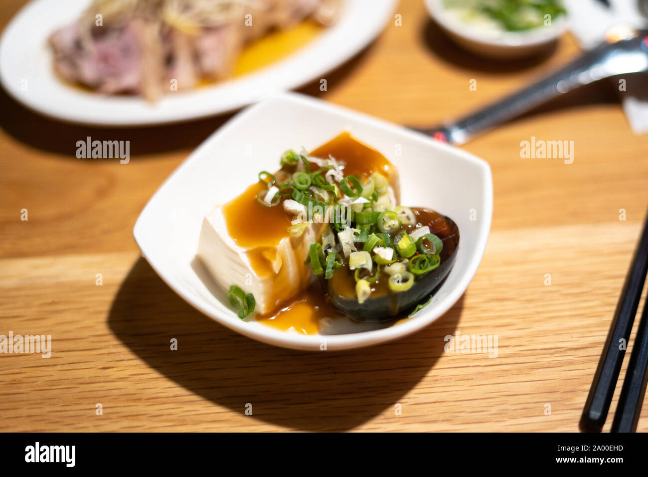 Weiße Schüssel mit silken Tofu und ein tausend Jahre altes Ei mit süßen Sojasoße und Frühlingszwiebeln auf einen hölzernen Tisch im Restaurant. 1000 Jahre altes Ei, 皮蛋 Stockfoto