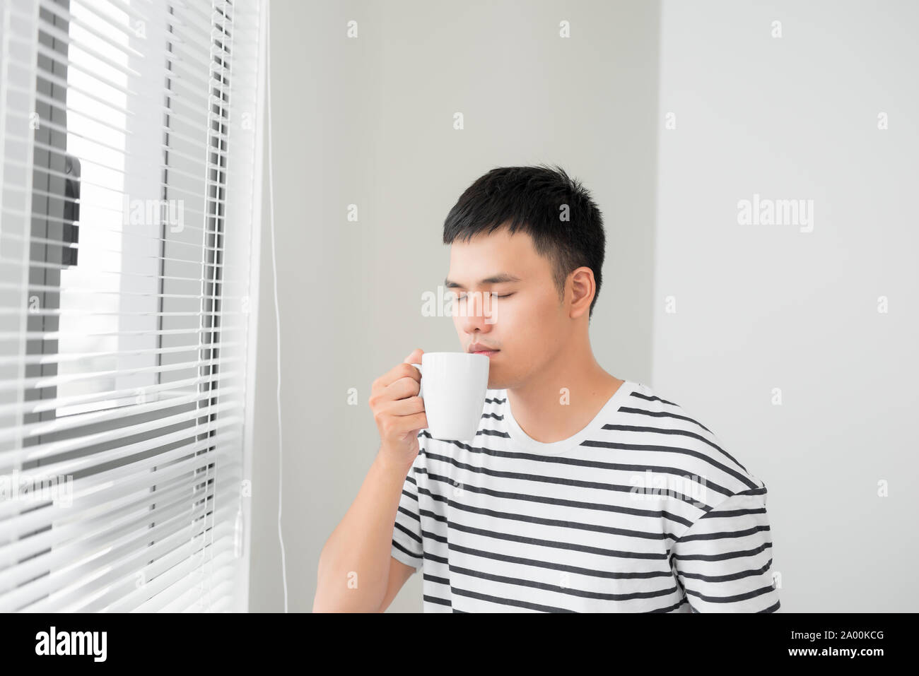 Junge schöne sorglose Mann in der Nähe von modernes Fenster in voller Länge genießen eine Tasse Kaffee, während außerhalb, guten Morgen suchen, Stockfoto