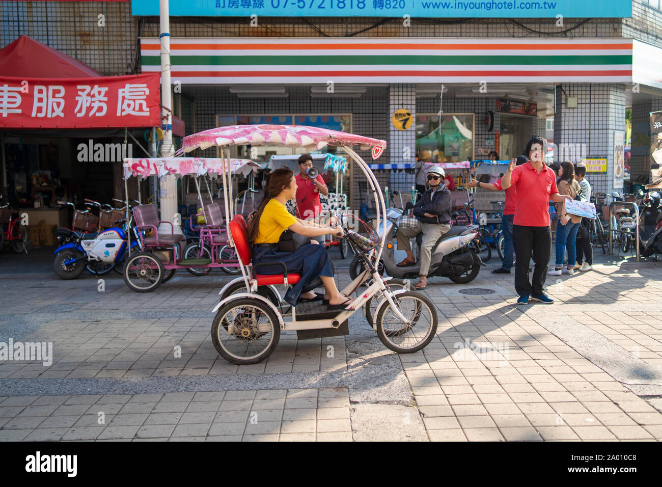 Kaohsiung, Taiwan: Zwei junge asiatische Frauen fahren ein Allradantrieb elektrische Fahrrad durch die Cijin Hauptstraße mit einem Seven Eleven (711) Stockfoto