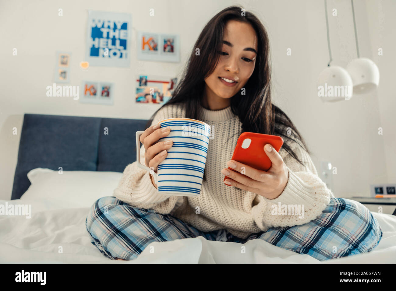 Lächelnde junge Dame in ihrem Bett sitzend mit einem Smartphone und einem großen Becher in der Hand Stockfoto