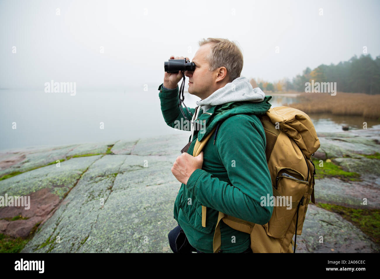 Reifer mann Finnland erkunden im Herbst, in Nebel Blick durch ein Fernglas. Wanderer mit grossen Rucksack auf bemoosten Felsen. Skandinavische Landschaft Stockfoto