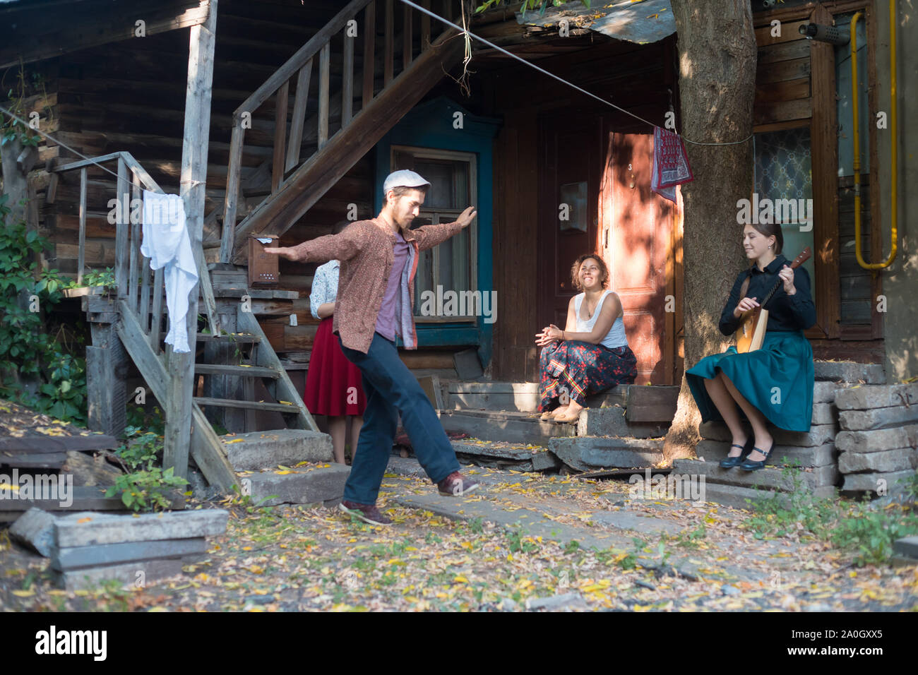 Ein Mann in weißen Hut tanzen russischen Volkstanz - Frauen sitzen auf der Treppe in der Nähe des Country House und ihm zuzusehen - eine Frau spielen Balalaika Stockfoto