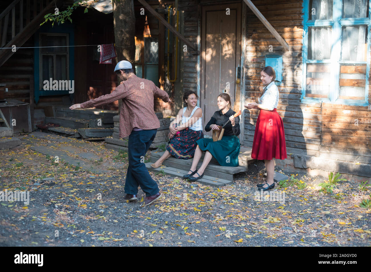 Ein Mann tanzen russischen Volkstanz - Frauen sitzen auf der Treppe in der Nähe des Country House und miteinander sprechen - eine Frau spielen Balalaika Stockfoto