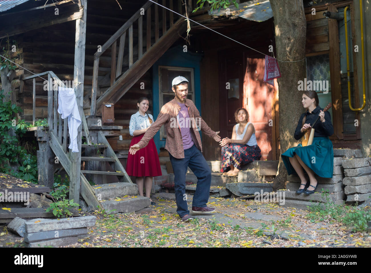 Ein bärtiger Mann tanzen russischen Volkstanz - Frauen sitzen auf der Treppe in der Nähe des Country House und ihm zuzusehen - eine Frau spielen Balalaika Stockfoto