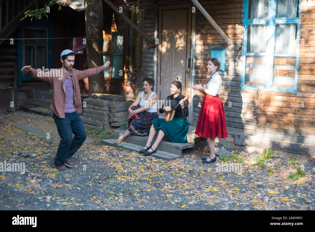 Ein Mann tanzen russischen Volkstanz - Frauen sitzen auf der Treppe in der Nähe des Country House ihm zuzusehen und Lächeln - eine Frau spielen Balalaika Stockfoto