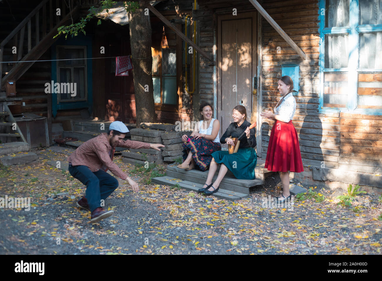 Ein Mann tanzen russischen Volkstanz - Frauen sitzen auf der Treppe in der Nähe des Country House ihm zuzusehen - Lachen und Lächeln - eine Frau spielen Balalaika Stockfoto