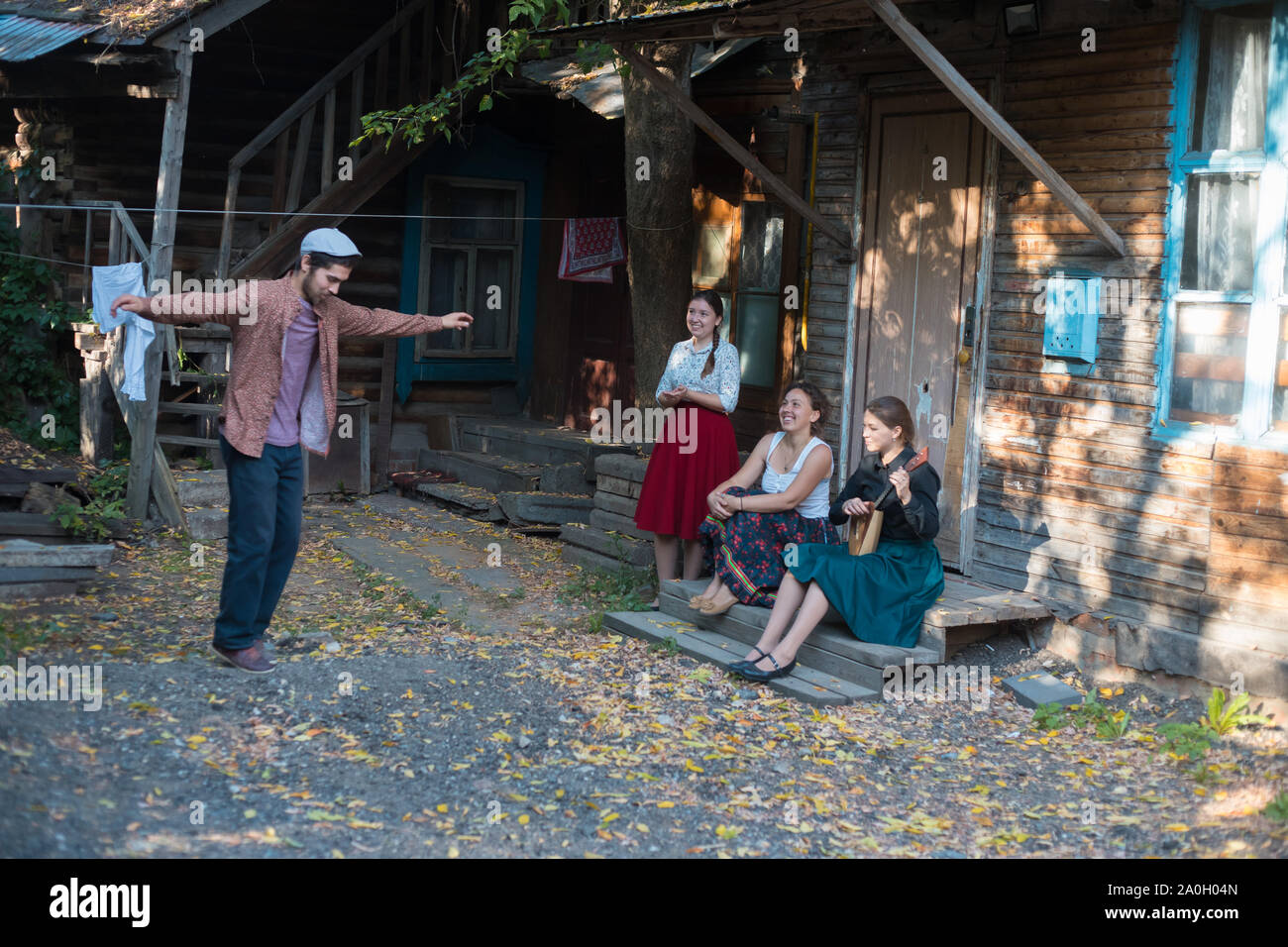 Ein Mann in weißen Hut tanzen russischen Volkstanz - Frauen sitzen auf der Treppe in der Nähe des Hauses ihn beobachtete und Lächeln - eine Frau spielen Balalaika Stockfoto