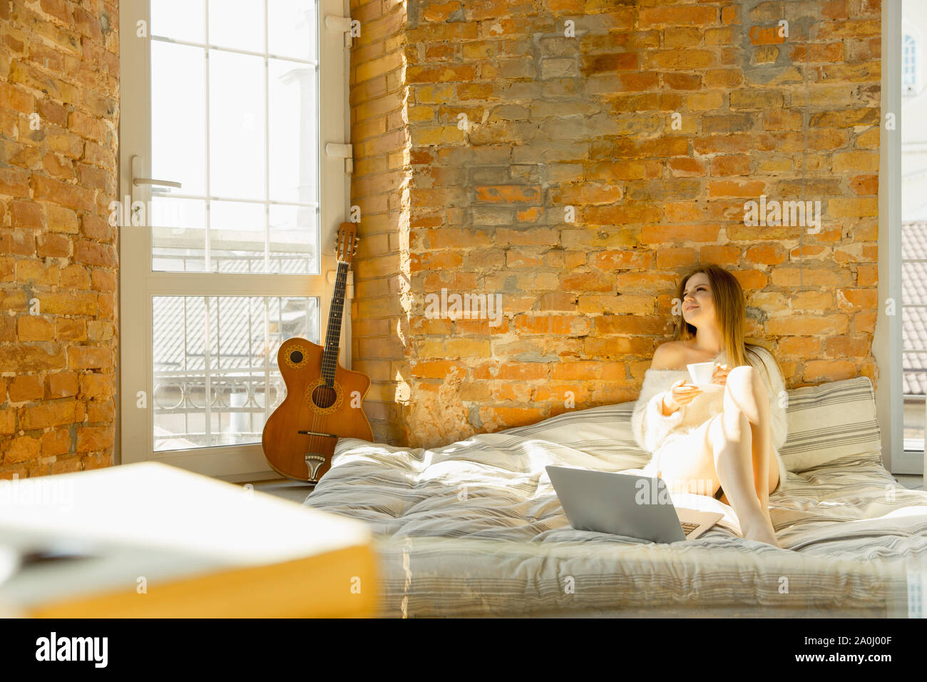 Entspannt zu Hause. Schöne junge Frau liegt auf Matratze mit warmen Sonnenlicht. Kaukasische blonde female model hat am Wochenende Zeit zum Ausruhen. Arbeiten an Ihrem Laptop und lächelnd. Die blog oder Surfen. Stockfoto