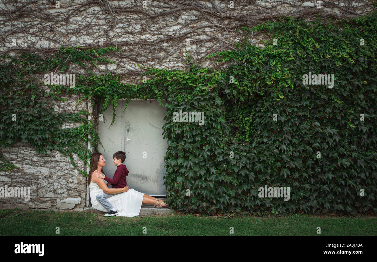 Mutter mit Sohn auf dem Schoß sitzen im Eingang eines Efeu bedeckten Wand. Stockfoto