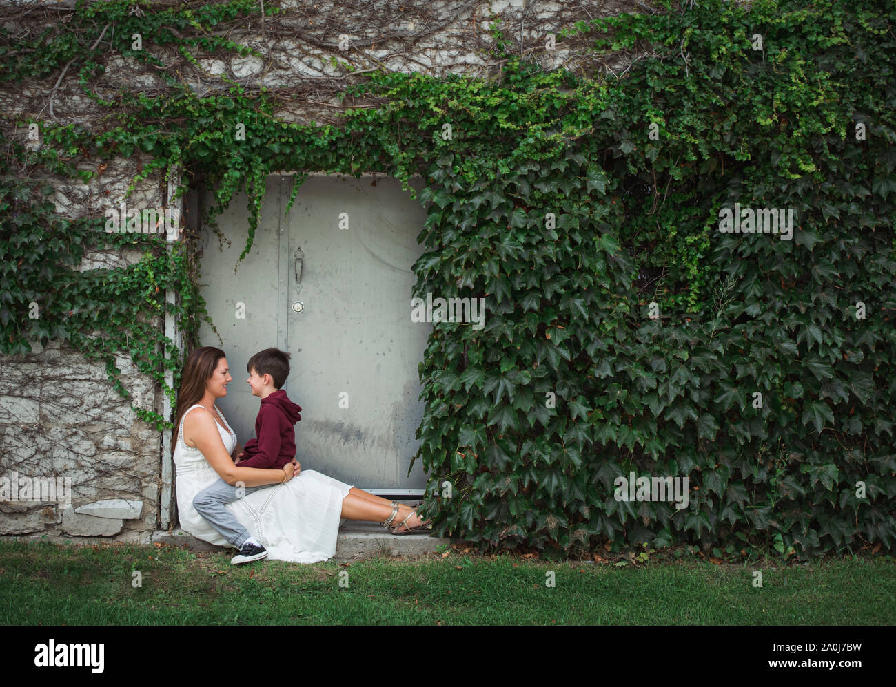 Mutter mit Sohn auf dem Schoß sitzen im Eingang eines Efeu bedeckten Wand. Stockfoto