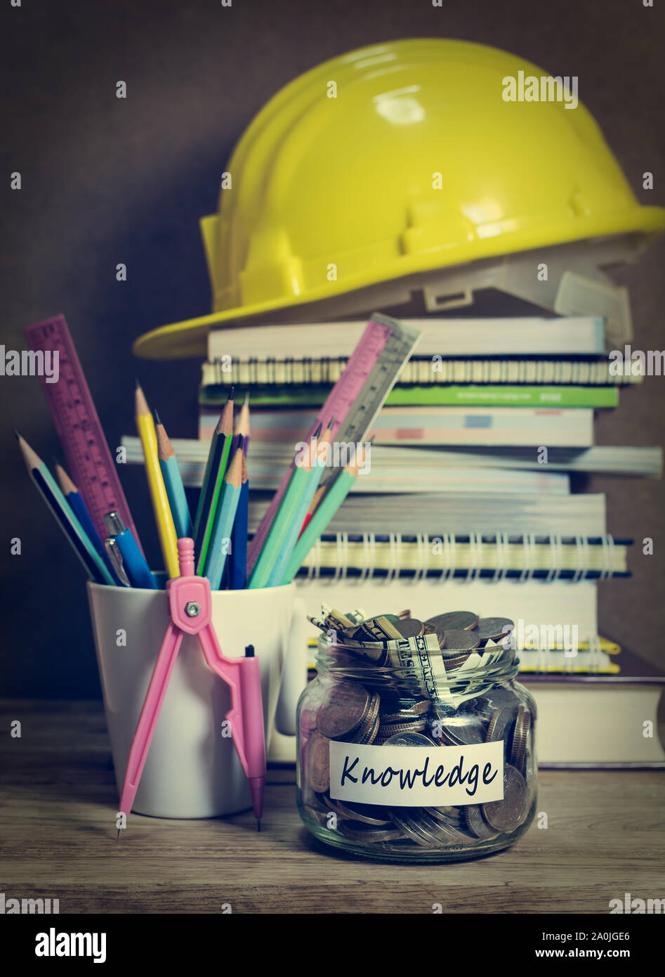 Geld sparen für Wissen im Glas Glas mit Haufen von Lehrbuch, Notepad und Bau Helm mit Schreiben Material auf hölzernen Schreibtisch Stockfoto
