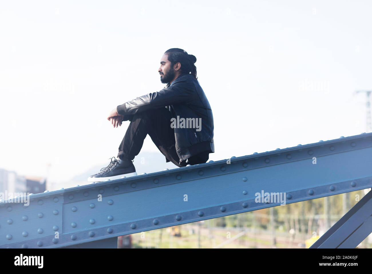 Junger Mann mit Zopf und Bart sittinging auf einer Stahlbrücke Stockfoto