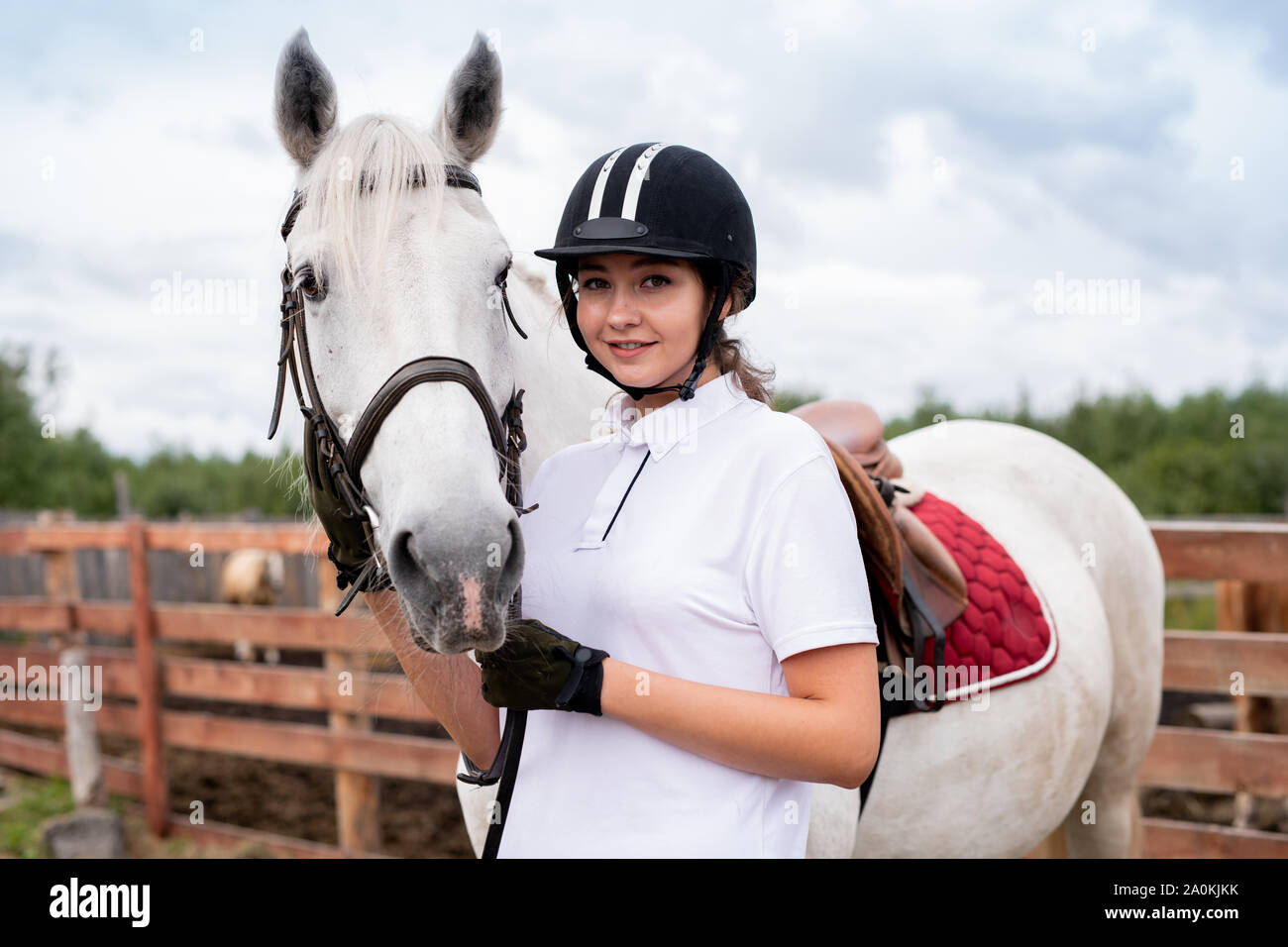 Junge lächelnde Frau in Pferdesport Outfit in der Nähe von weißen Rennpferd stehend Stockfoto