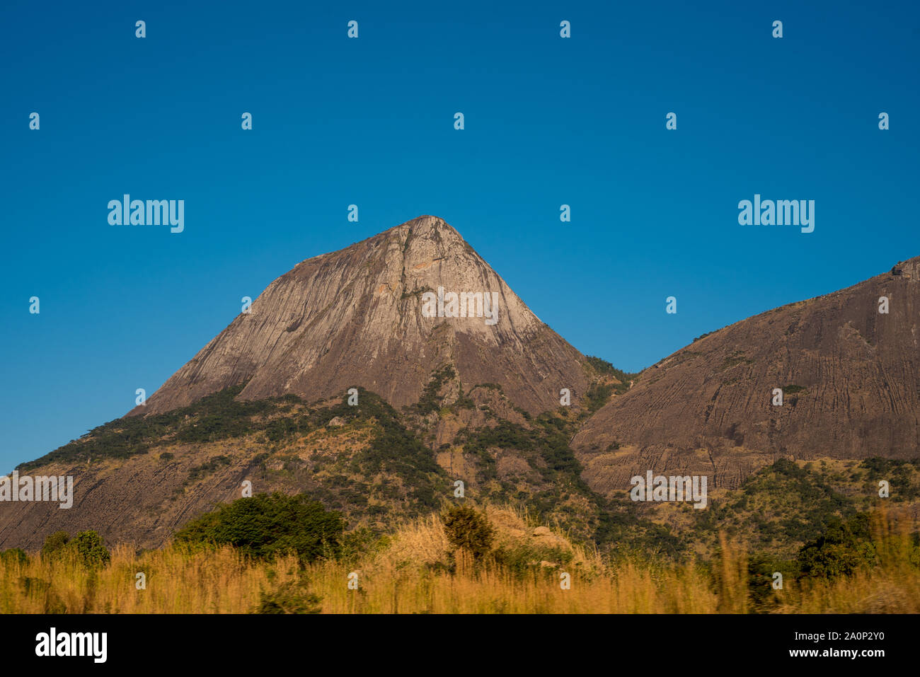 Die felsigen und konisch geformte Berg erhebt sich steil aus dem grasbewachsenen Ebenen unter einem klaren blauen Himmel im ländlichen Mosambik, Afrika Stockfoto