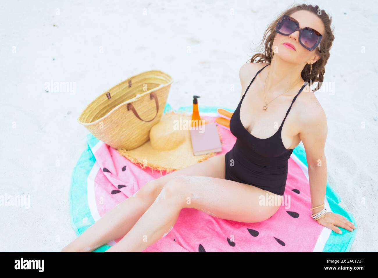 Obere Ansicht von trendigen 40 Jahre alte Frau in der Sonnenbrille mit langen Locken im eleganten schwarzen Badeanzug an einem weißen Strand sonnenbaden während der Sitzung auf Runden Stockfoto