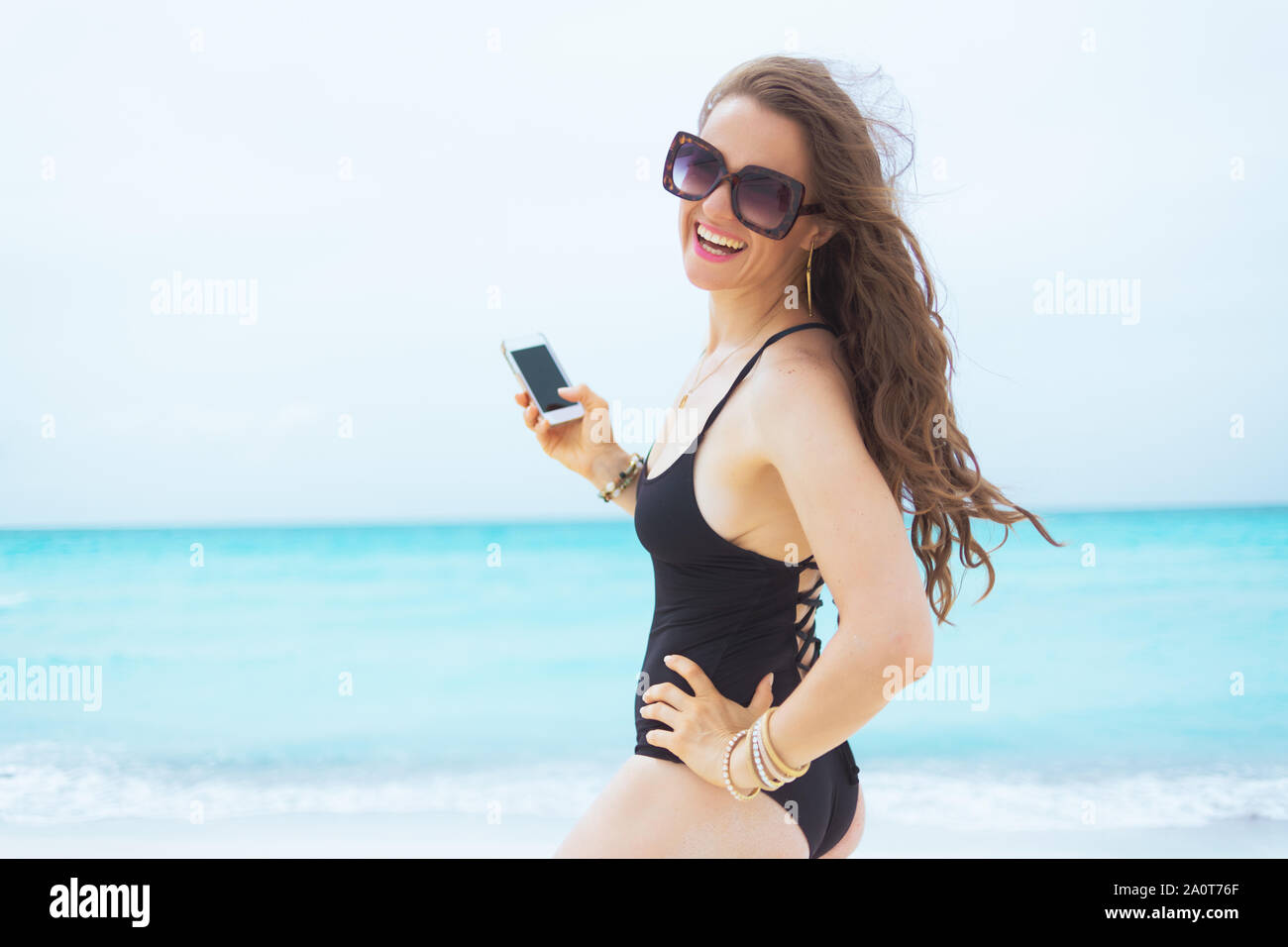 Portrait von lächelnden elegante 40-jährige Frau in Sonnenbrille mit Smartphone Verwenden von Multimedia Programmen an einem weißen Strand. Stockfoto