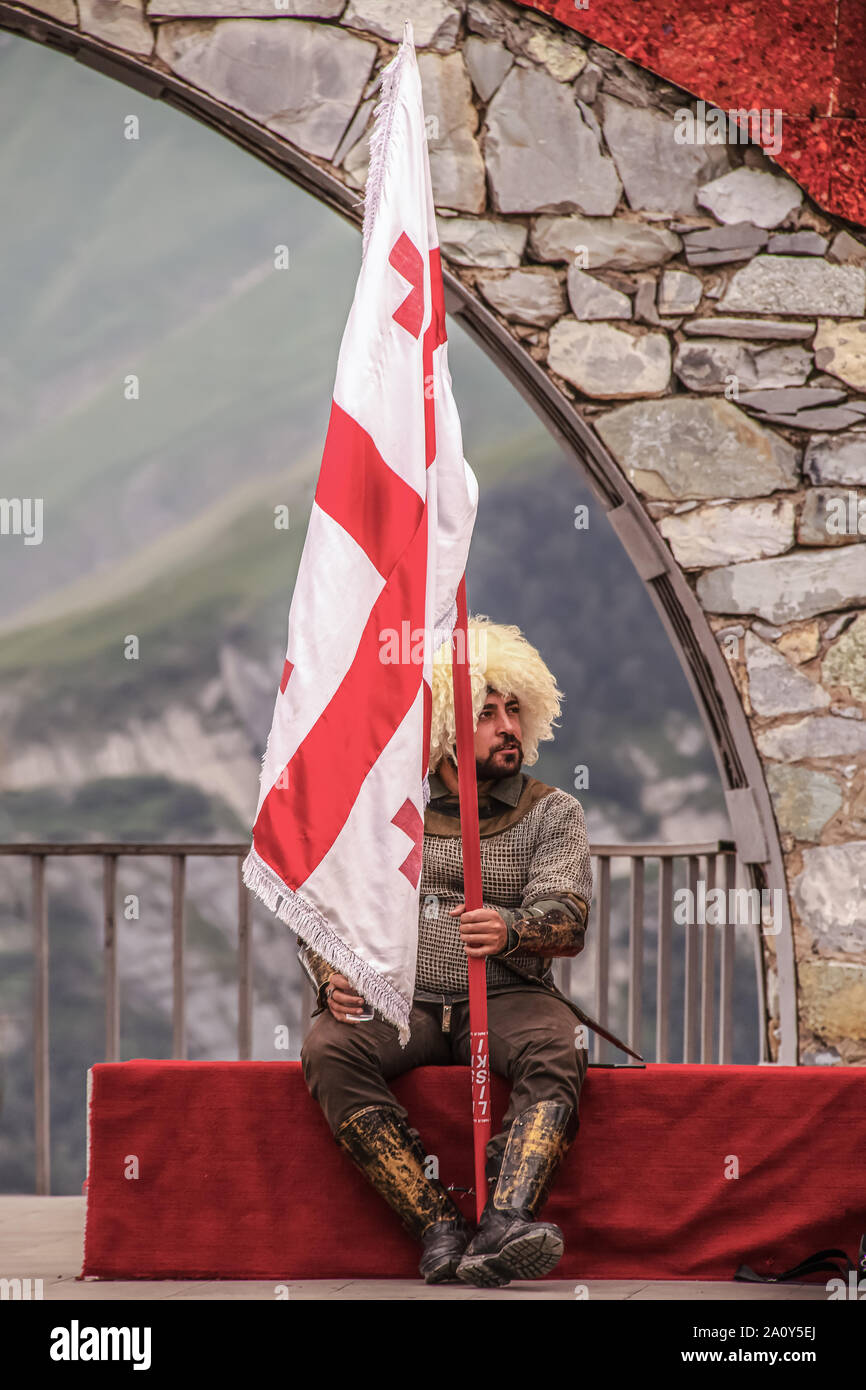 6-16-2019 Military Highway Georgien - Mann in der traditionellen georgischen Kleidung mit Pelzmütze holding Flag gekleidet sitzt in Russland - Georgien Friendship Monument Stockfoto