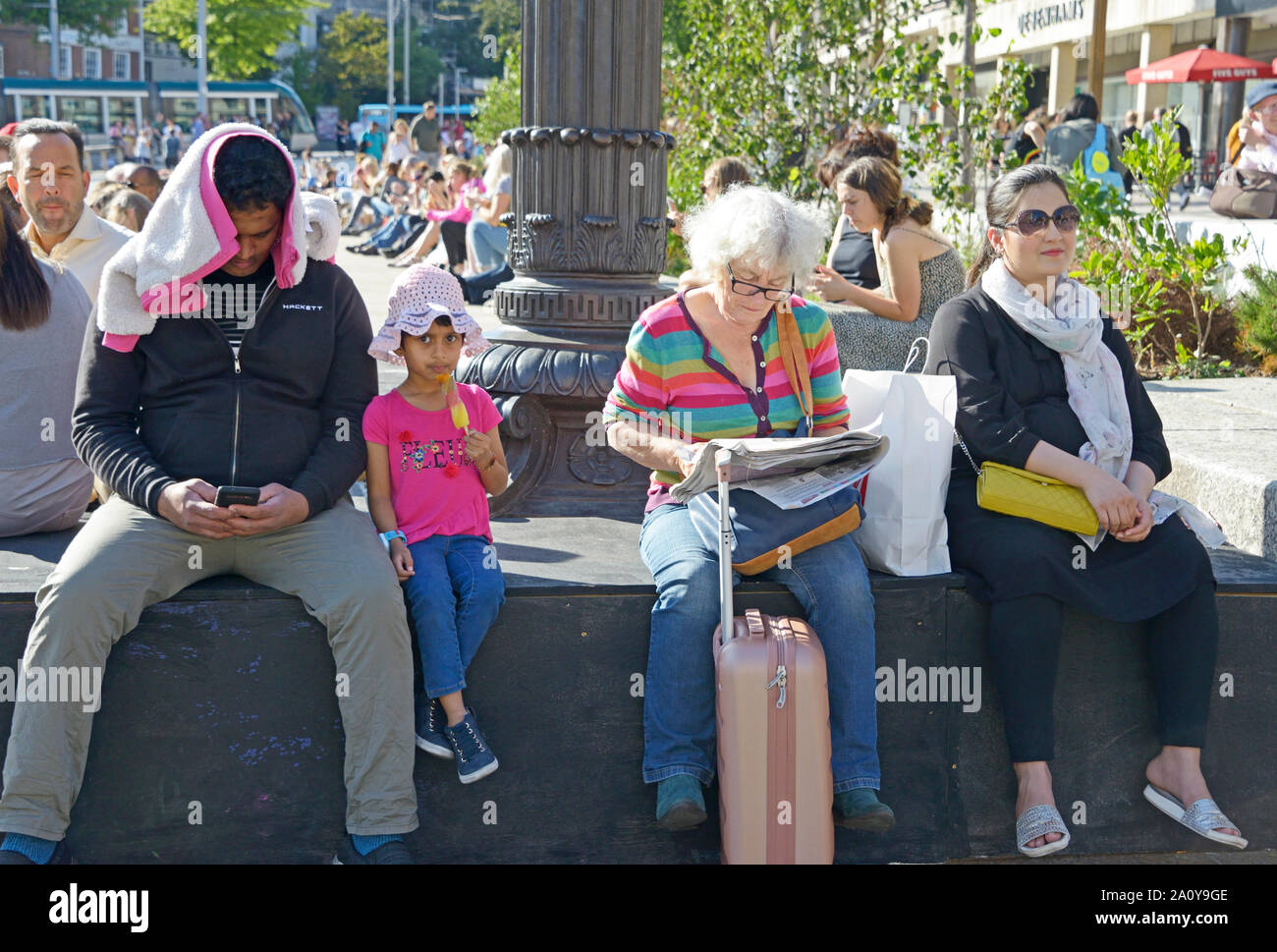 Leute sitzen auf einer Mauer. Gemischte Gruppe. Mann am Telefon, Kind, alte Dame & eine andere. Stockfoto