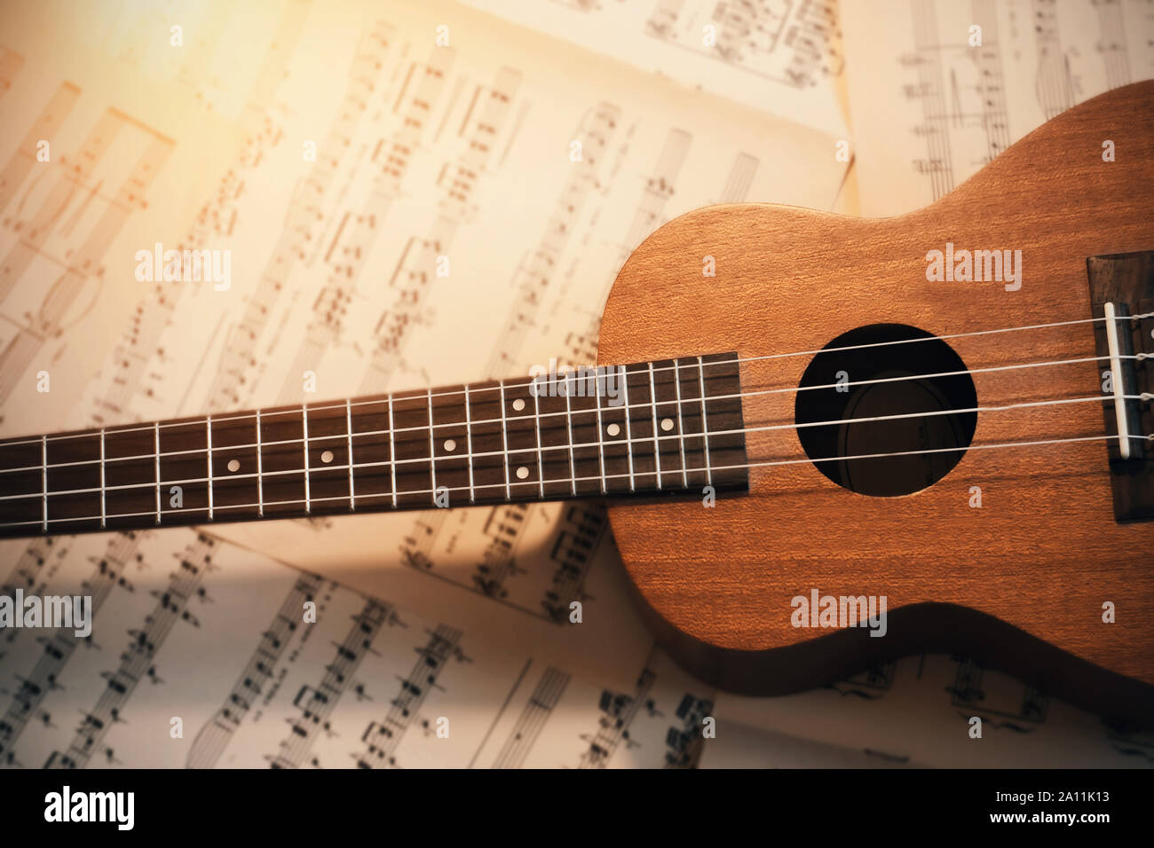 Eine kleine hölzerne Ukulele mit Nylonsaiten liegt auf verstreute Blätter der Noten, durch das Licht leuchtet Stockfoto