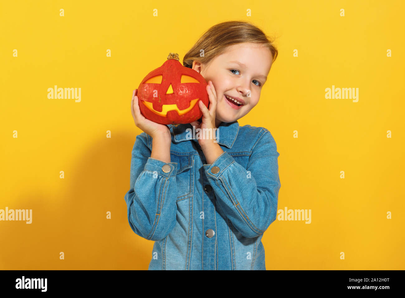 Portrait von ein fröhliches kleines Mädchen in einem Jeanshemd. Ein Kind hält einen Kürbis jack Lantern auf gelbem Hintergrund. Halloween Konzept. Stockfoto