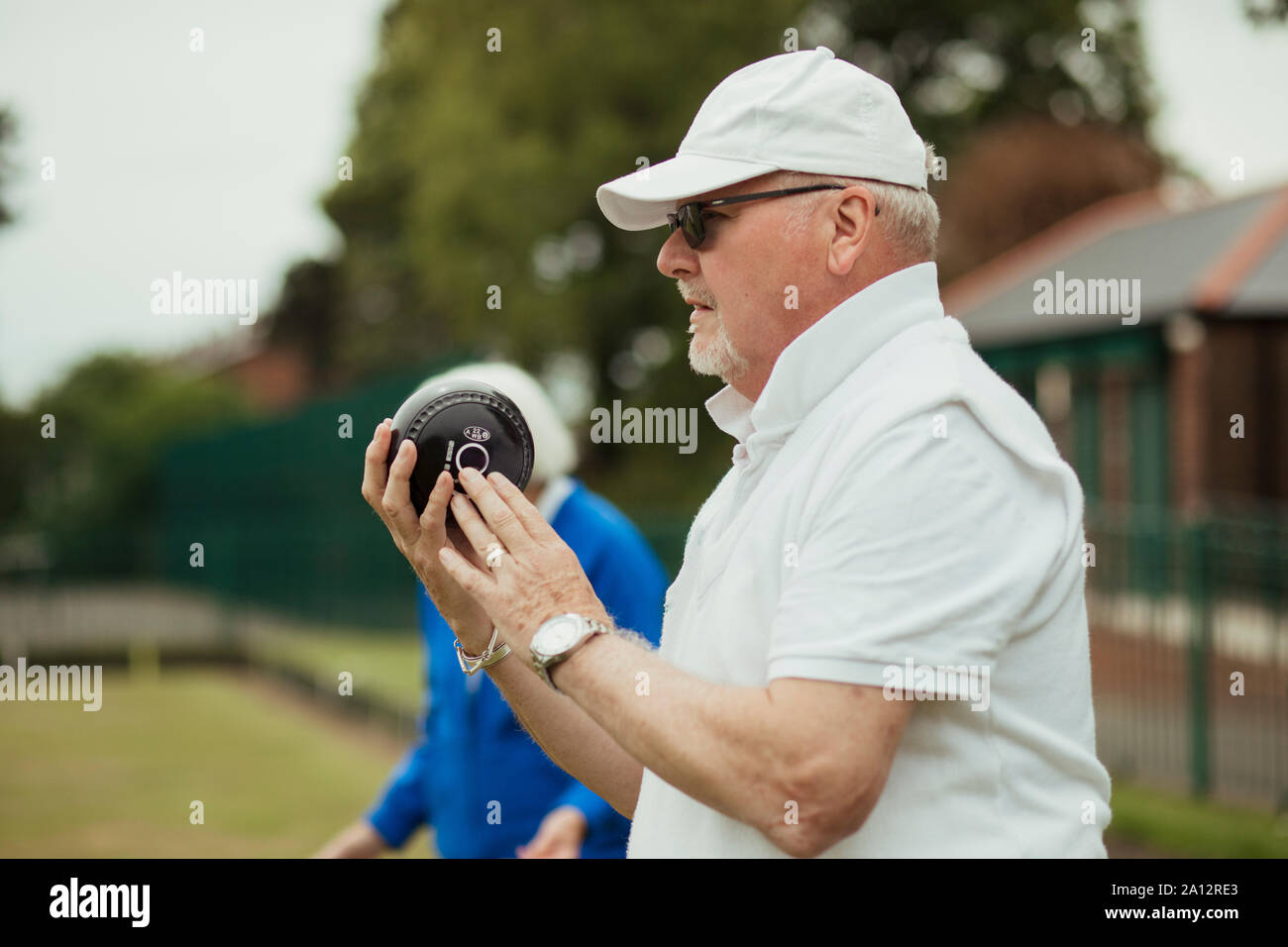 Eine Nahaufnahme von einem fokussierten älterer Mann hält eine Boccia, bereit, seinen Schoß zu nehmen. Er trägt eine weiße Kappe und Sonnenbrille. Stockfoto