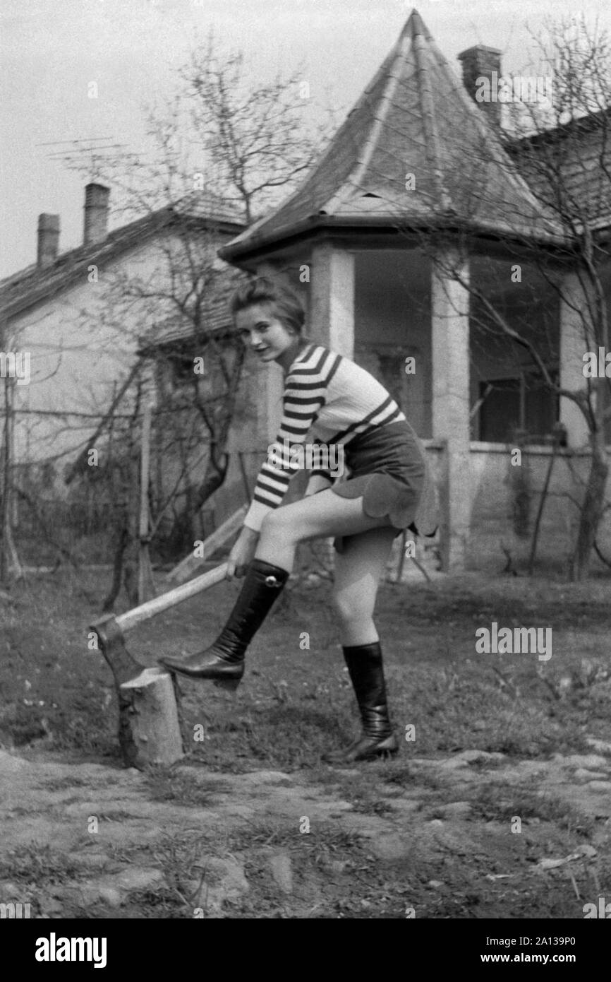 Junge Frau, die lange schwarze Stiefel mit Mini Rock im Garten mit Axt und Baumstumpf Hackklotz 1970 s Ungarn Stockfoto