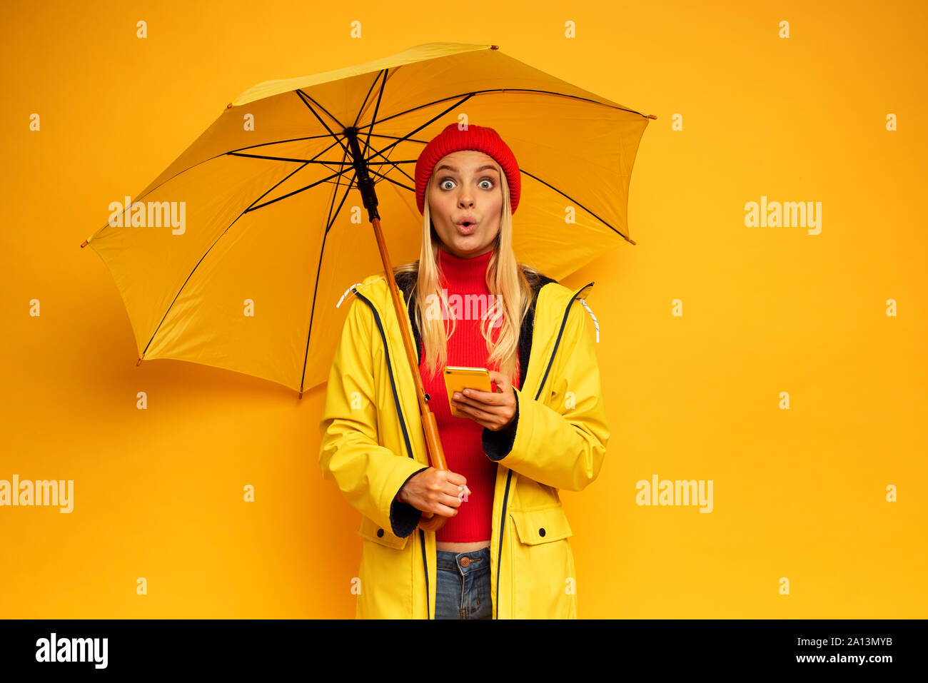 Mädchen mit Smartphone und Sonnenschirm auf gelben Hintergrund für das Wetter überrascht Stockfoto