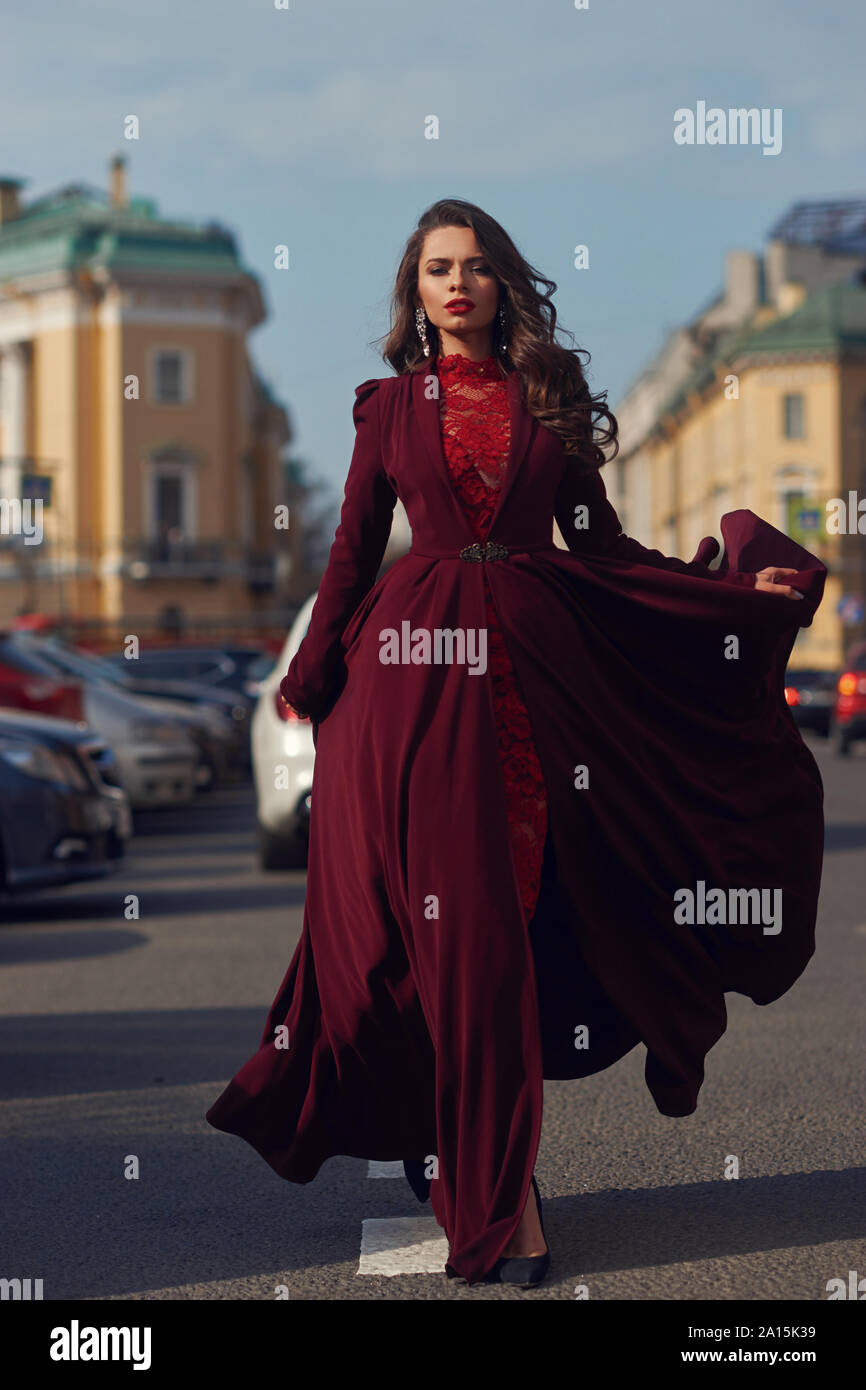 Junge schöne atemberaubende Frau in Rot luxus Abendkleid mit fliegenden  Saum Walking City Straße an einem sonnigen Abend. Elegante Dame mit Make-up  und gewellt b Stockfotografie - Alamy