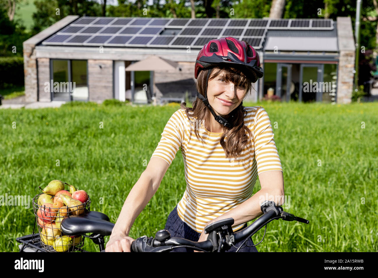 Porträt der lächelnde Frau mit Fahrrad und biologischem Obst auf einer Wiese vor einem Haus Stockfoto