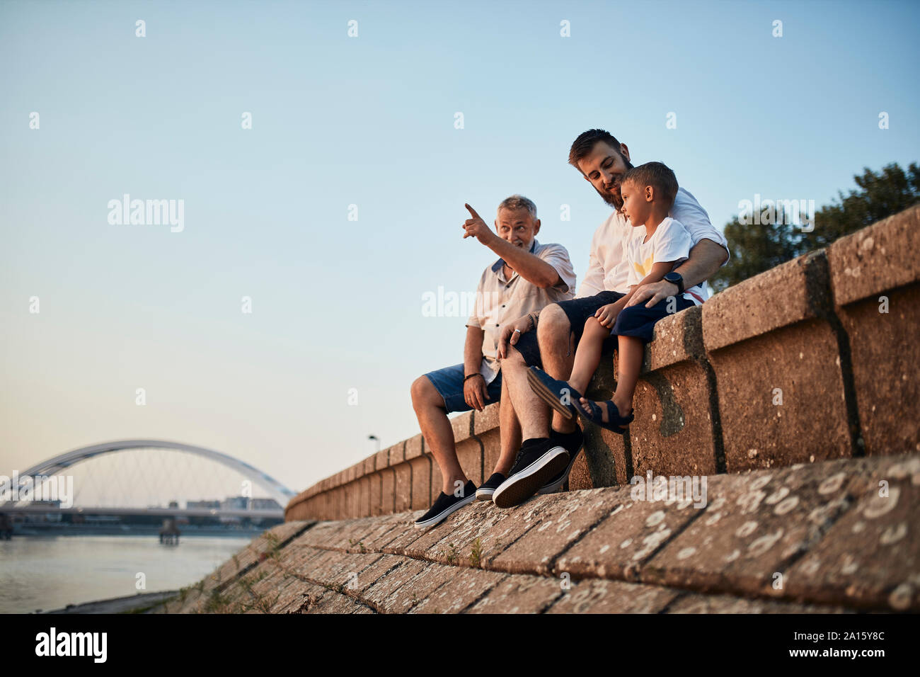 Großvater, Vater und Sohn sitzen auf einer Mauer am Flußufer Stockfoto