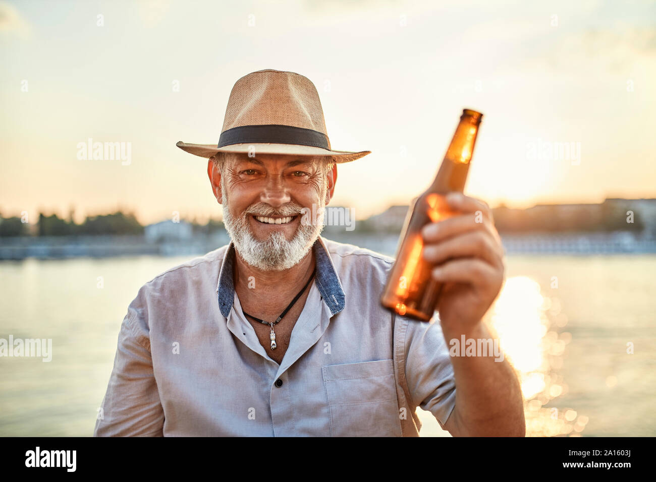 Portrait Of Happy älterer Mann ein Bier trinken im Riverside bei Sonnenuntergang Stockfoto