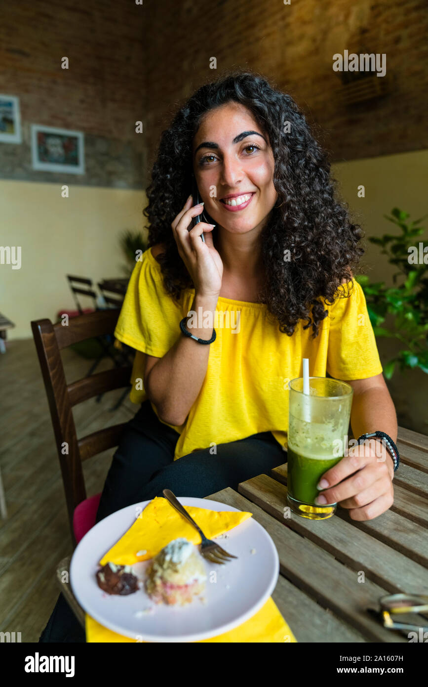 Portrait von lächelnden jungen Frau auf Handy am Tisch sitzend mit einem Smoothie Stockfoto