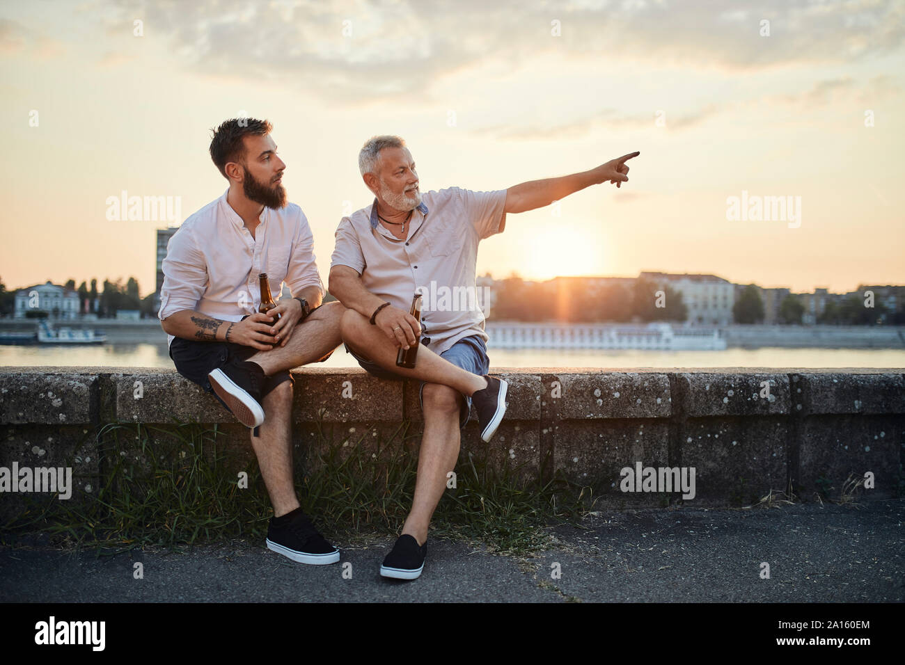 Vater und Sohn sitzen auf einer Mauer an der Riverside bei Sonnenuntergang ein Bier trinken Stockfoto