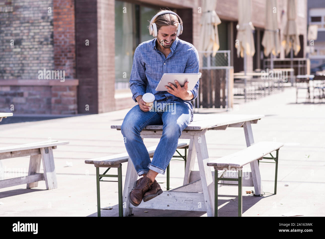 Junge Mann sitzt am Tisch in einem Biergarten mit Kopfhörern und Tablet Stockfoto