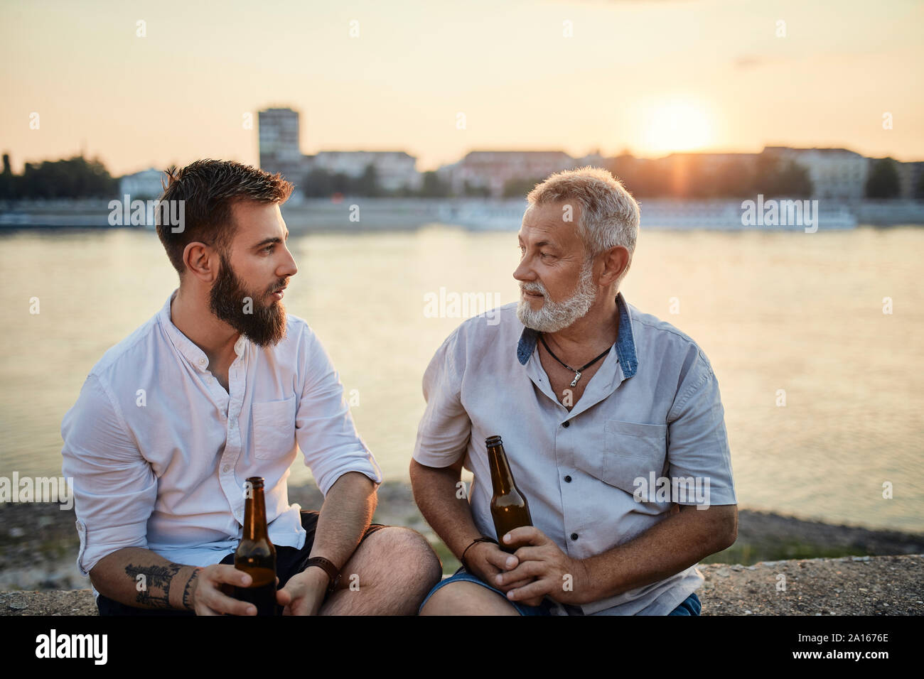 Vater und Sohn sitzen auf einer Mauer an der Riverside bei Sonnenuntergang ein Bier trinken Stockfoto