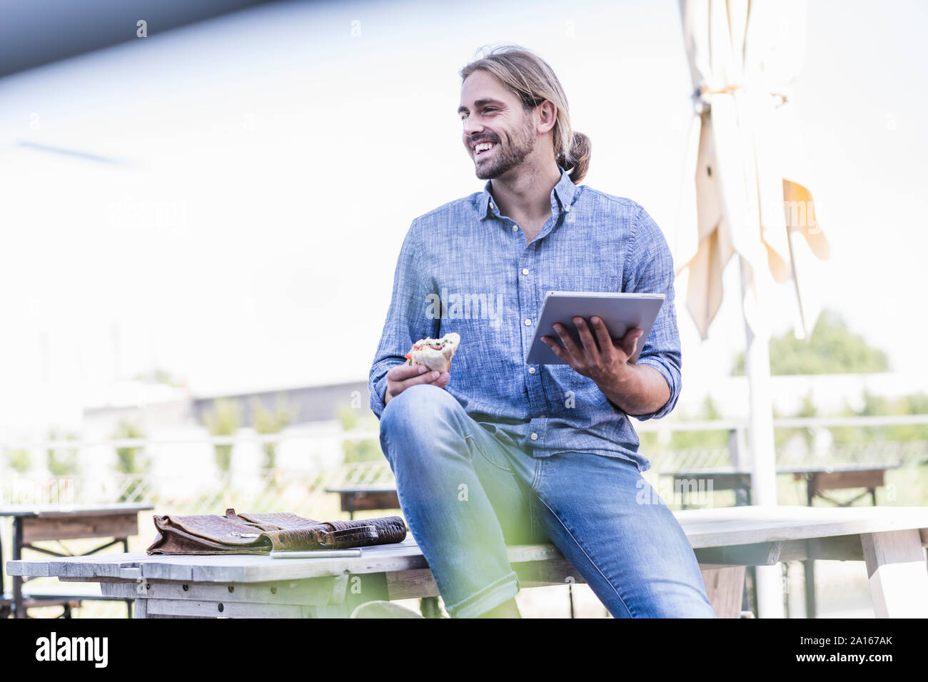 Junge Mann sitzt am Tisch in einem Biergarten mit Tablet und Sandwich Stockfoto