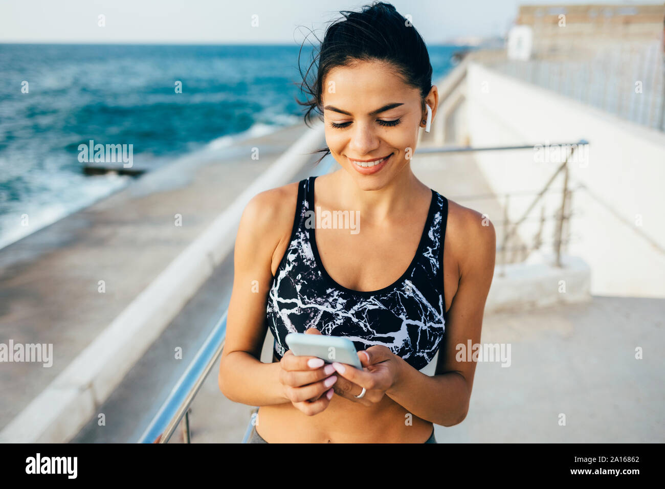 Frau mit Smartphone und In-ear während des Trainings, sitzend auf einem Pier Stockfoto