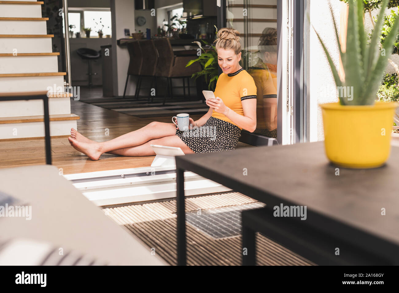 Entspannt Frau an der Terrassentür zu Hause mit Handy sitzen, Kaffee Tasse und Tablet Stockfoto