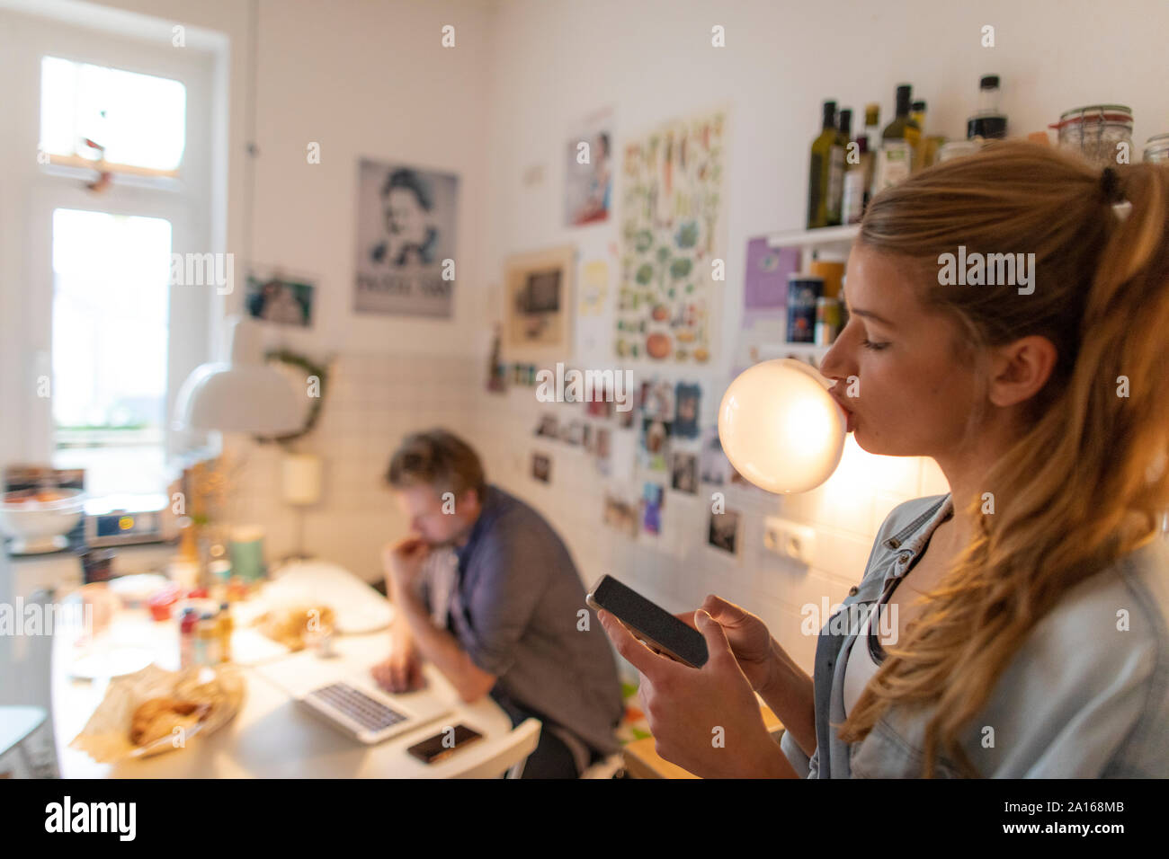 Junge Frau in der Küche zu Hause Kaugummi bubble mit Partner im Hintergrund Stockfoto