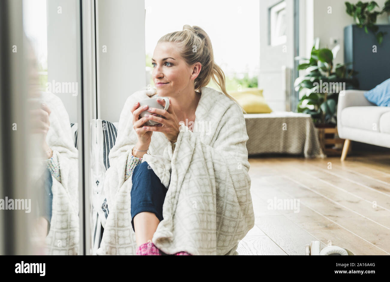 Frau eingehüllt in eine Decke am Fenster sitzen zu Hause Kaffee trinken Stockfoto