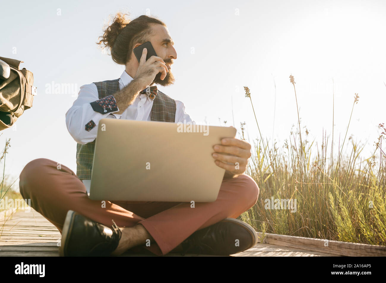 Gut gekleideter Mann sitzt auf einem Holzsteg in der Landschaft mit Laptop und Mobiltelefon Stockfoto