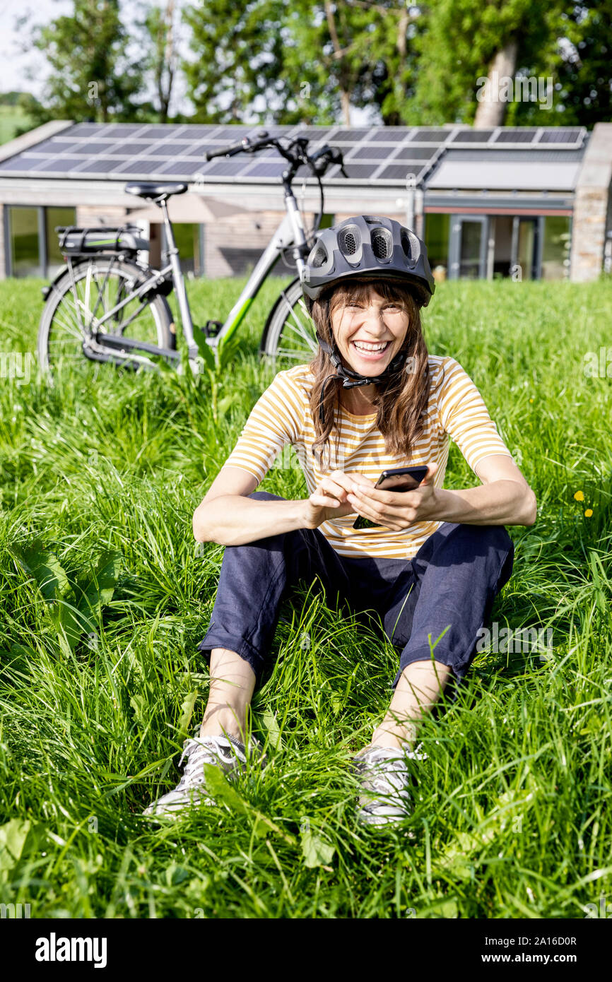 Lachende Frau mit Fahrrad mit Handy auf einer Wiese vor einem Haus Stockfoto